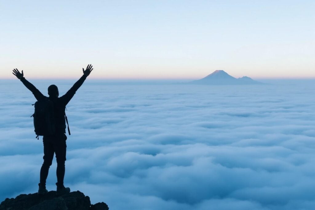 Silhouetted hiker above clouds at sunrise, mountain distant